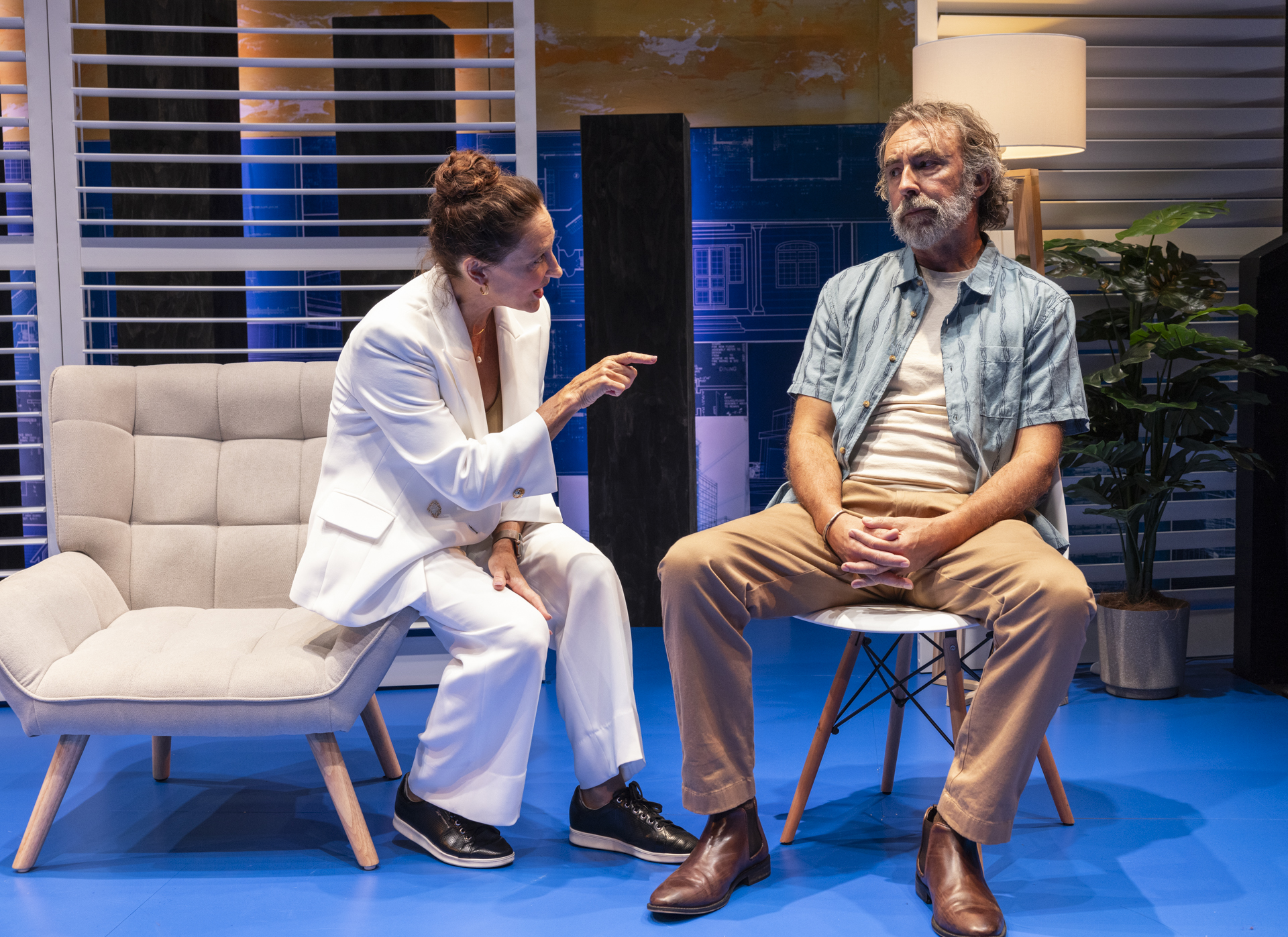 A woman in a white suit gestures seriously towards a seated older man in casual clothes. They are on a blue stage set with modern chairs, plants, and blinds, suggesting a tense conversation in a contemporary setting.