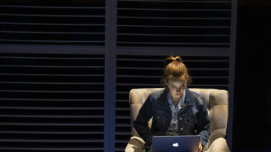 A young person sits on an armchair using a laptop, illuminated by a beam of light. A rucksack is on the floor beside them, and the room features large, dark window blinds in the background.
