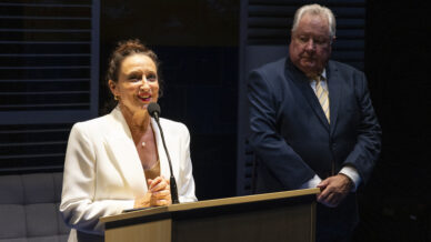 A woman in a white blazer speaks at a lectern with a microphone, smiling, while a man in a dark suit and striped tie stands behind her, watching, in a dimly lit room.