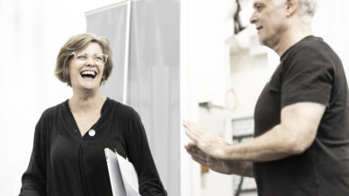 A woman holding papers and smiling warmly stands next to a man in conversation. Both appear engaged and are dressed in black, against a bright, indoor background.