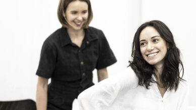 Two women indoors, one in a black outfit standing and smiling in the background, and another in a white shirt sitting in the foreground, looking to the side and smiling. The atmosphere appears friendly and relaxed.