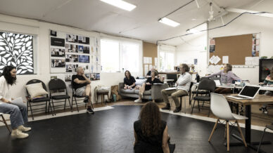 Several people sit apart in a spacious, well-lit room with white walls, noticeboards, and a black floor. Some are in chairs, one sits on the floor, and they appear to be having a casual group discussion or rehearsal.