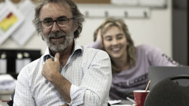 A man with glasses and a beard smiles and sits in the foreground, whilst a woman in the background, also smiling, leans on a table in an office or classroom setting. Papers and folders are visible on the wall.