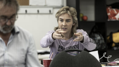 A woman with short blonde hair wearing a light purple shirt gestures with both hands whilst speaking in a classroom or meeting room. A man sits in the foreground, out of focus. Papers and bags are visible in the background.