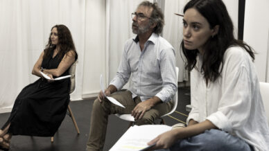 Three people sit on chairs indoors holding papers, appearing focused and attentive. White curtains hang in the background, and the setting suggests a rehearsal or meeting.