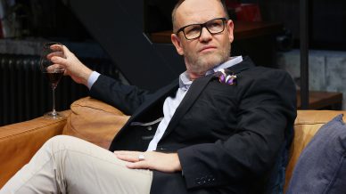 A man wearing glasses and a black blazer sits on a tan sofa, holding a glass of wine. He appears contemplative, with one arm resting on the back of the sofa and his legs crossed.