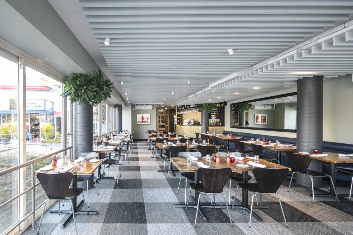 Modern restaurant interior with large windows, wooden tables set for dining, dark chairs, chequer-patterned carpet, and ceiling with white beams. Bright and airy ambience with plants and a view outside.