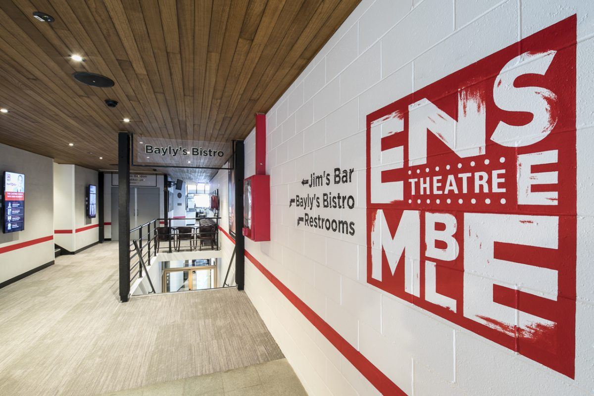 A modern corridor with timber ceiling, white walls, and red accents leads to Bayly’s Bistro, Jim’s Bar, and toilets. A large red-and-white Ensemble Theatre logo is painted on the wall.
