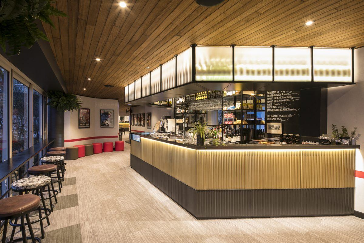 Modern bar with illuminated counter, high stools, and plants. Large windows line one side, whilst framed posters hang on the walls. The ceiling is wood-panelled, and the space features warm, ambient lighting.