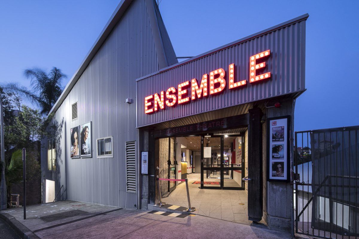 Exterior view of Ensemble Theatre at dusk, featuring a corrugated metal façade and a large, red, illuminated ENSEMBLE sign above the entrance. Posters are displayed near the entrance and on the building's side wall.