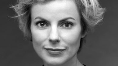 Black and white portrait of a woman with short, tousled blonde hair, wearing a dark top, looking directly at the camera with a neutral expression against a plain background.