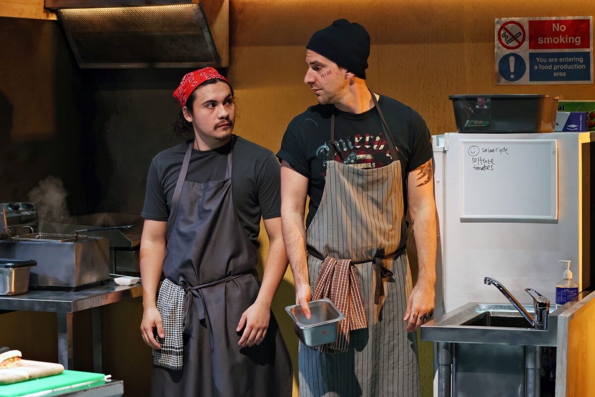 Two men wearing aprons stand in a commercial kitchen. One wears a red bandana, the other a black beanie. They look at each other seriously, surrounded by kitchen equipment and a “No Smoking” sign on the wall.