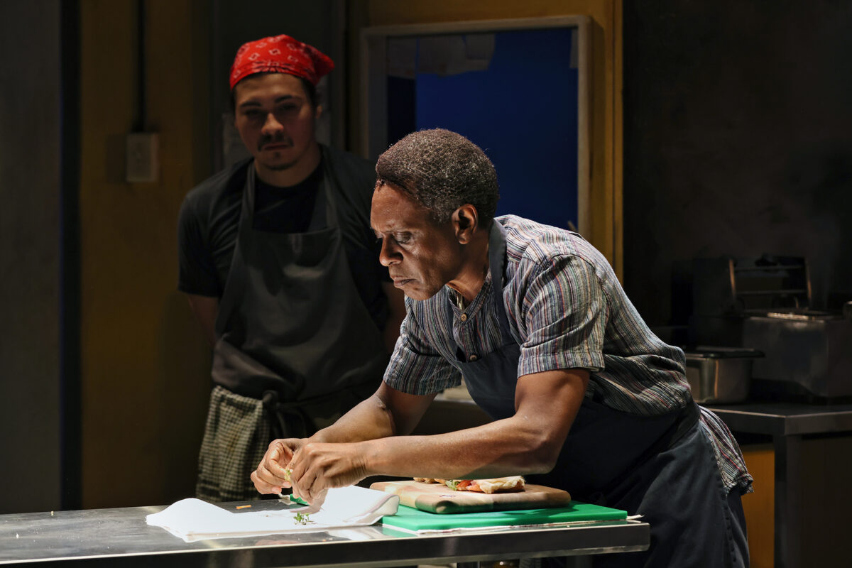 A man in an apron prepares food intently at a kitchen worktop while another man in a red bandana and dark clothing stands in the background, watching. The setting appears to be a professional kitchen.