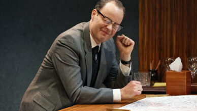 A man in a grey suit and glasses sits at a wooden desk, smiling with his eyes closed and raising his fist cheerfully. Papers, a glass, and desk items are on the table in front of him.