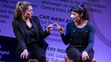Two women sit on stools, facing each other and pointing fingers. One wears a black suit, the other denim dungarees and a plaster on her nose. The background features large, partially visible text from classified adverts.
