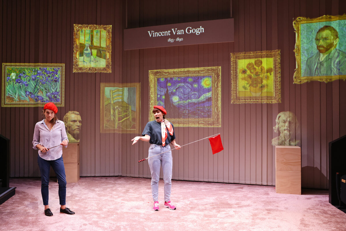 Two performers in red berets stand onstage, each holding a red flag, with Vincent van Gogh’s name and several of his famous paintings projected behind them. Busts of artists sit on plinths at the sides of the stage.
