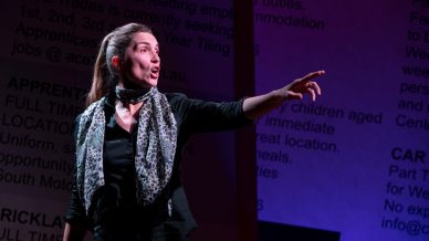 A woman with long hair tied back, wearing a leopard print scarf and black shirt, stands under stage lighting with her mouth open and arm outstretched, appearing to perform dramatically. Text is projected on the wall behind her.