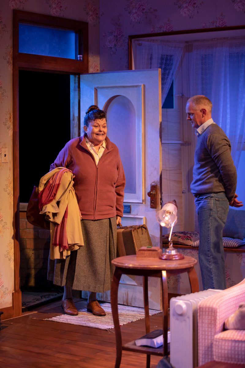 An older woman holding a coat and suitcase stands in an open doorway, smiling at a man inside a cosy, warmly lit room with floral wallpaper and vintage décor. The man looks at her with his hands behind his back.