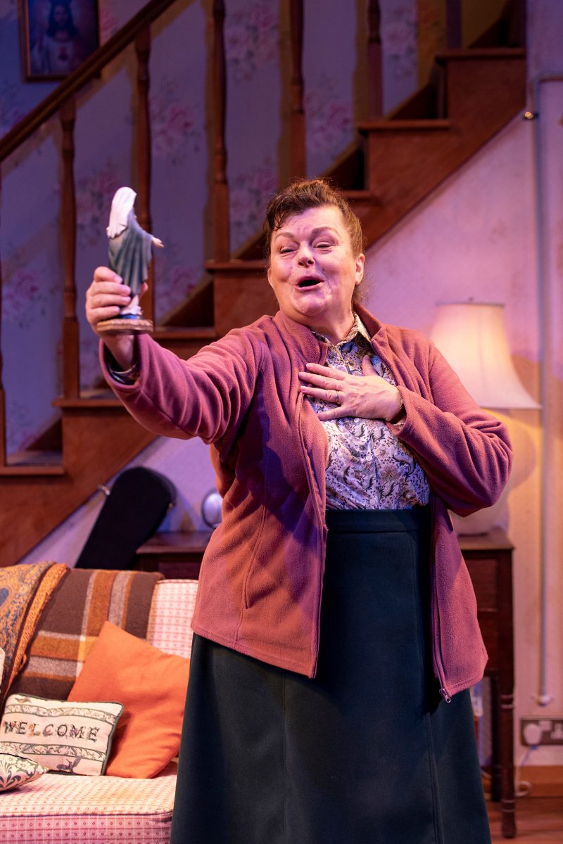 A woman stands in a cosy living room, holding up a small statuette with one hand and touching her chest with the other, appearing joyful or moved. A staircase and warmly lit lamp are visible in the background.