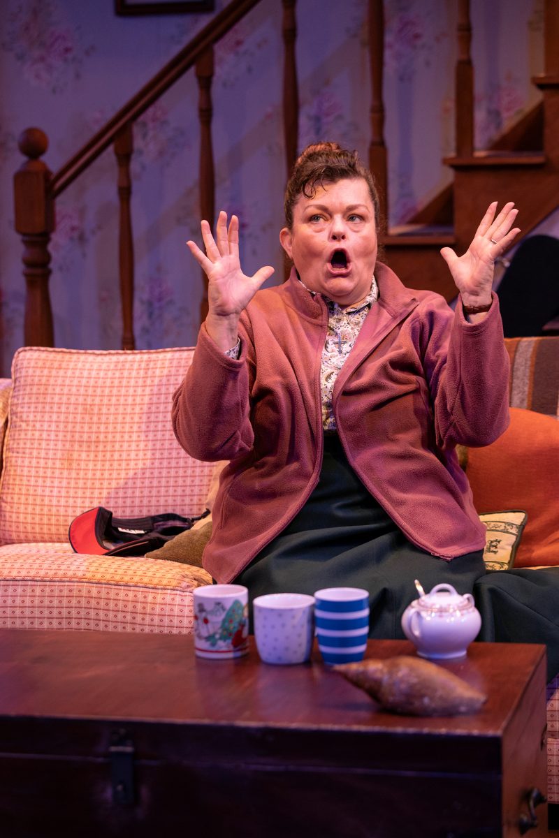 A woman sits on a sofa with a surprised expression, raising both hands in the air. Three mugs, a teapot, and a pastry are on the coffee table in front of her. A staircase and floral wallpaper are in the background.