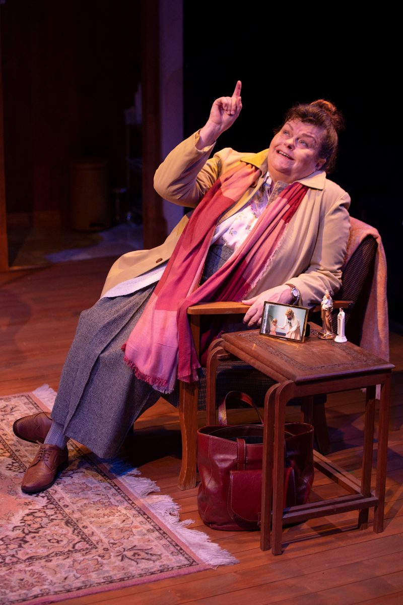 An older woman sits on a chair at a small wooden table, smiling and gesturing upward with one finger raised. She wears a beige coat, pink scarf, grey skirt, and brogues. The table holds framed photos and small figurines.