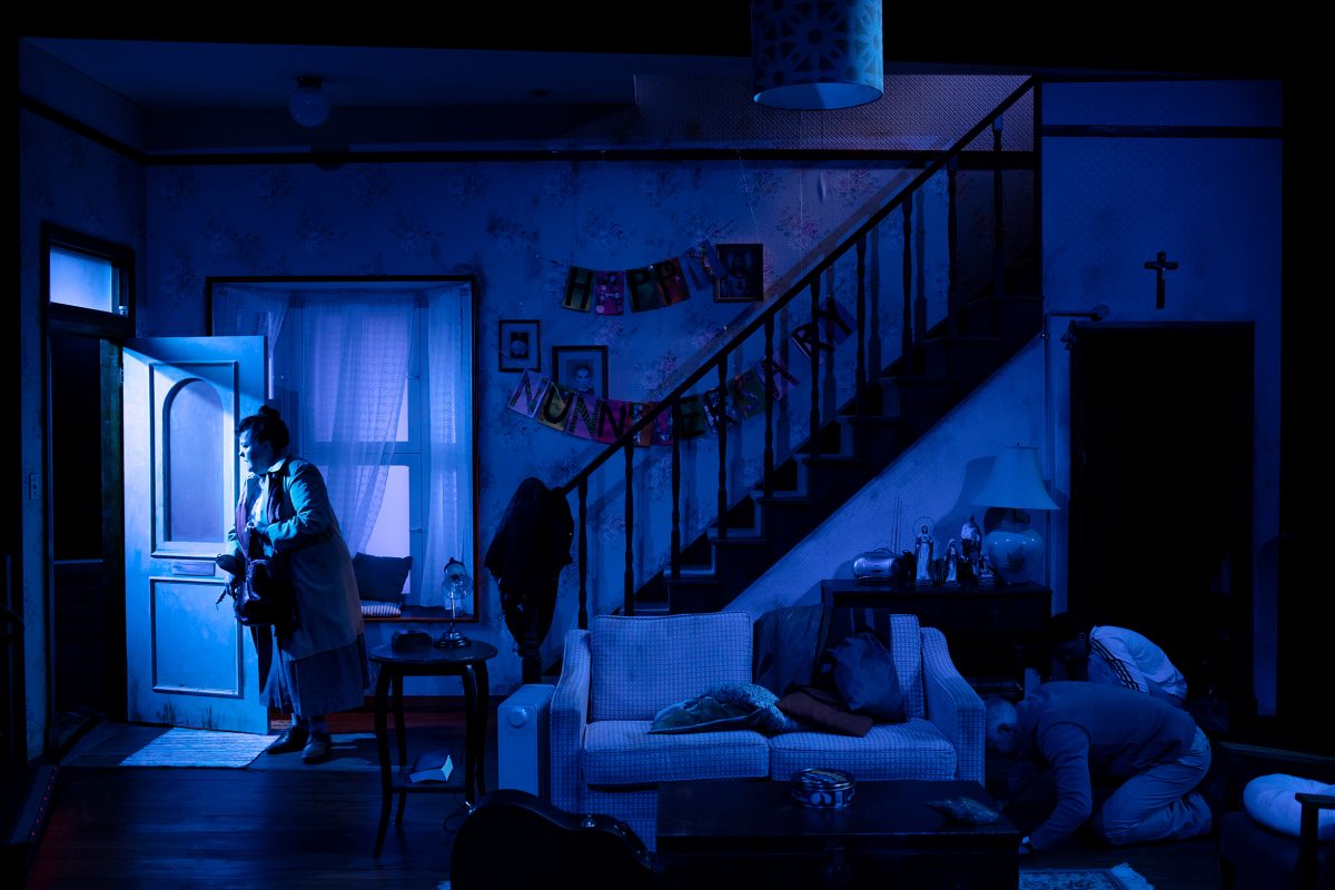 A dimly lit, blue-toned living room scene shows a person standing by an open door, half outside, while another person kneels on the floor near a sofa. A banner reading HAPPY ANNIVERSARY hangs on the staircase.