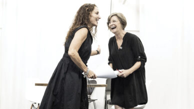 Two women in black dresses stand smiling and laughing together indoors, holding papers, in front of a white background with white chairs behind them.