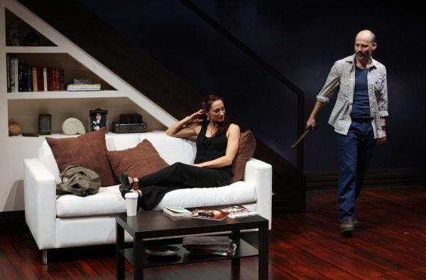 A woman sits on a white sofa with brown cushions, looking at a man standing nearby holding a wooden object. The setting appears to be a modern sitting room with shelves under a staircase.