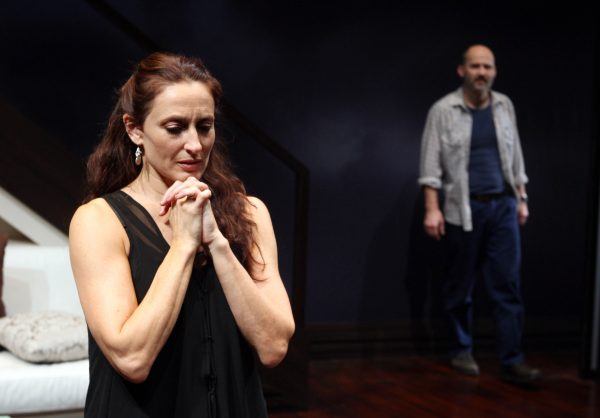 A woman stands in the foreground with her hands clasped, looking down pensively, whilst a man stands in the background, partially blurred, in a dimly lit room with dark walls and wooden floor.