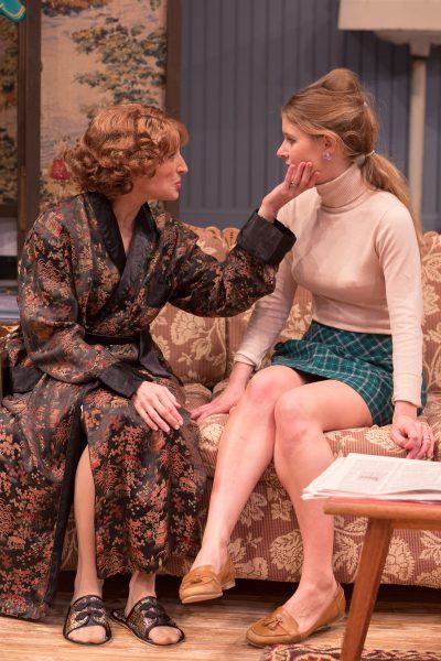 Two women sit closely on a patterned sofa. The older woman, in a floral dressing gown, gently touches the younger woman’s face. The younger woman wears a polo-neck jumper and checked skirt. They appear to be having a serious conversation.