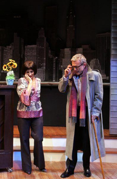 A woman in a floral top and trousers stands beside a man wearing a long coat, scarf, and glasses, leaning on a walking stick. Both appear to be acting emotionally on a stage with a cityscape backdrop.