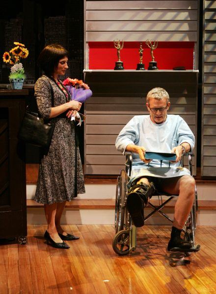A woman holding flowers stands beside a man in a wheelchair who is reading a document. Behind them, two trophies are displayed on a shelf against a wall with horizontal panels.