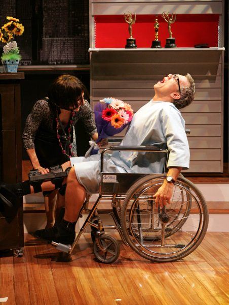 A person in a wig and costume stands next to a man in a wheelchair holding colourful flowers, with his head thrown back dramatically. Trophies are displayed on a shelf in the background.