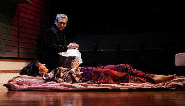 A woman in colourful clothing lies on a blanket on a stage, looking up at a man in dark clothes kneeling beside her, holding a white towel. Empty chairs are visible in the background.