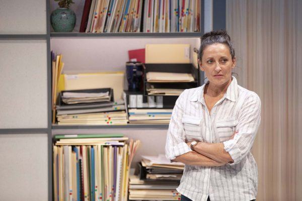 A woman with dark hair tied back, wearing a light striped shirt, stands with arms folded in front of a shelf filled with folders, files, and a green vase. She appears thoughtful or concerned.