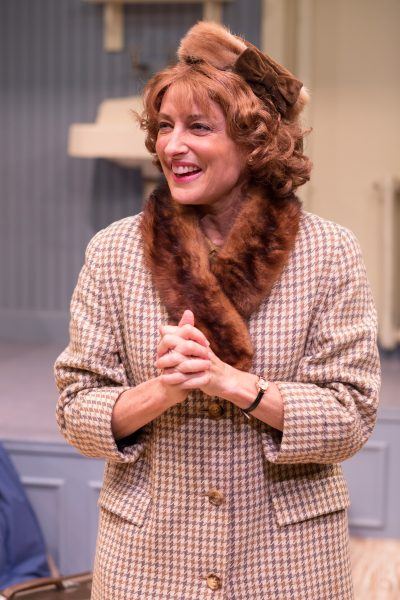 A smiling woman in a vintage houndstooth coat with a fur collar and matching brown hat stands indoors, hands clasped in front of her, appearing cheerful and engaged.