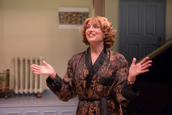 A woman with curly, shoulder-length hair smiles and gestures with open hands. She is wearing a patterned dressing gown and stands indoors near a staircase and a blue door.