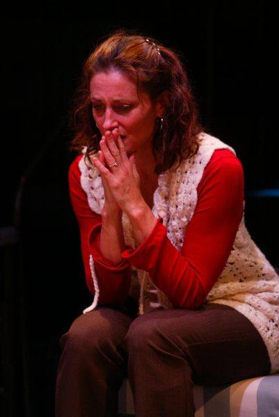 A woman with wavy brown hair sits with her hands covering her mouth, appearing anxious or deep in thought. She wears a red long-sleeved shirt and a white crocheted waistcoat. The background is dark and out of focus.