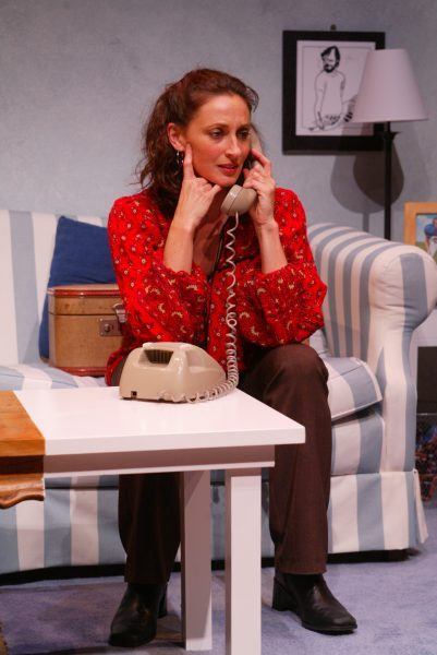 A woman with brown hair sits on a striped sofa, holding a corded telephone to her ear and face. She wears a red patterned blouse and brown trousers, with a neutral expression, in a cosy living room setting.