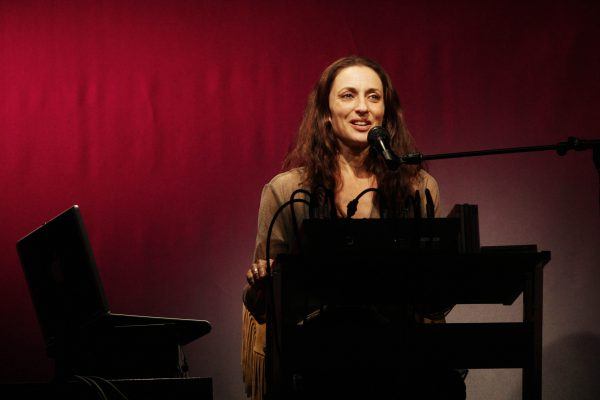 A woman with long brown hair speaks into a microphone whilst sitting at a desk with electronic equipment and a laptop, against a red and grey gradient background.