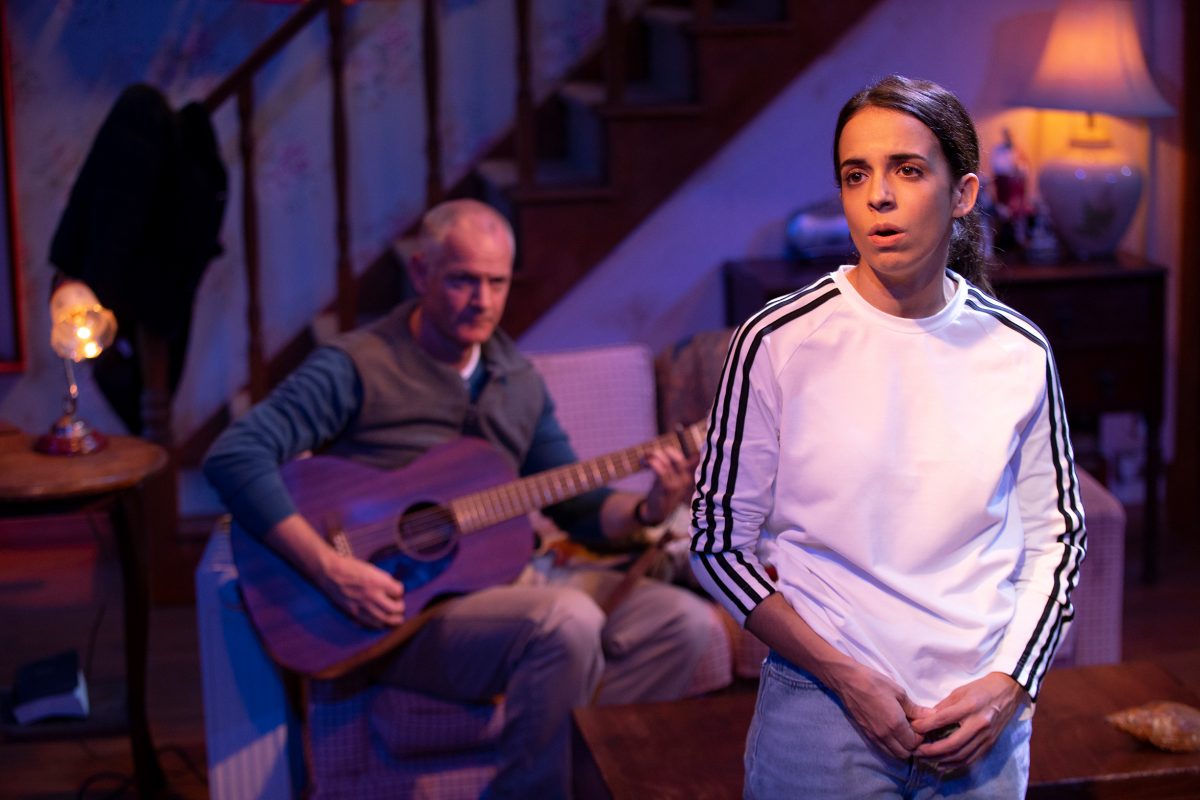 A woman in a white shirt stands in the foreground looking serious, while an older man in the background sits on a sofa playing a purple guitar. The scene takes place in a warmly lit living room with stairs and a lamp visible.