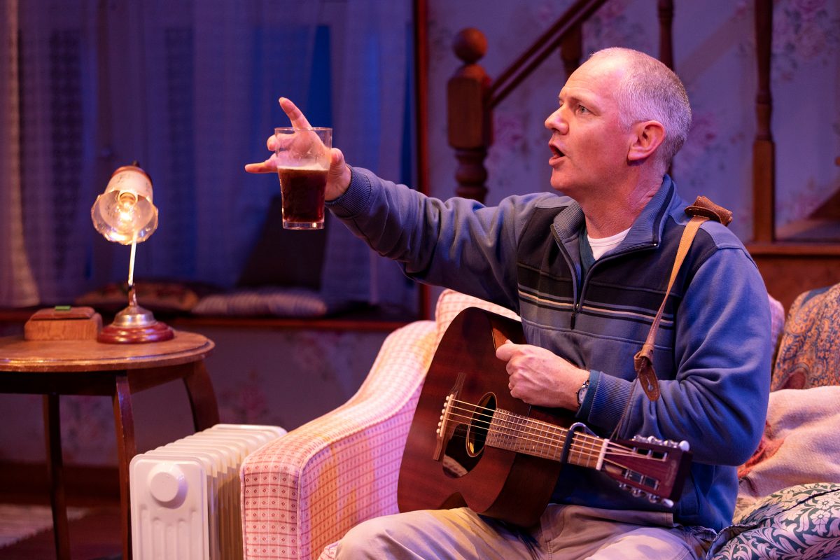 A man sits on a sofa holding a guitar and raising a glass, gesturing as if speaking or singing. A lit lamp, small table, and radiator are beside him, with a staircase and curtains in the background.