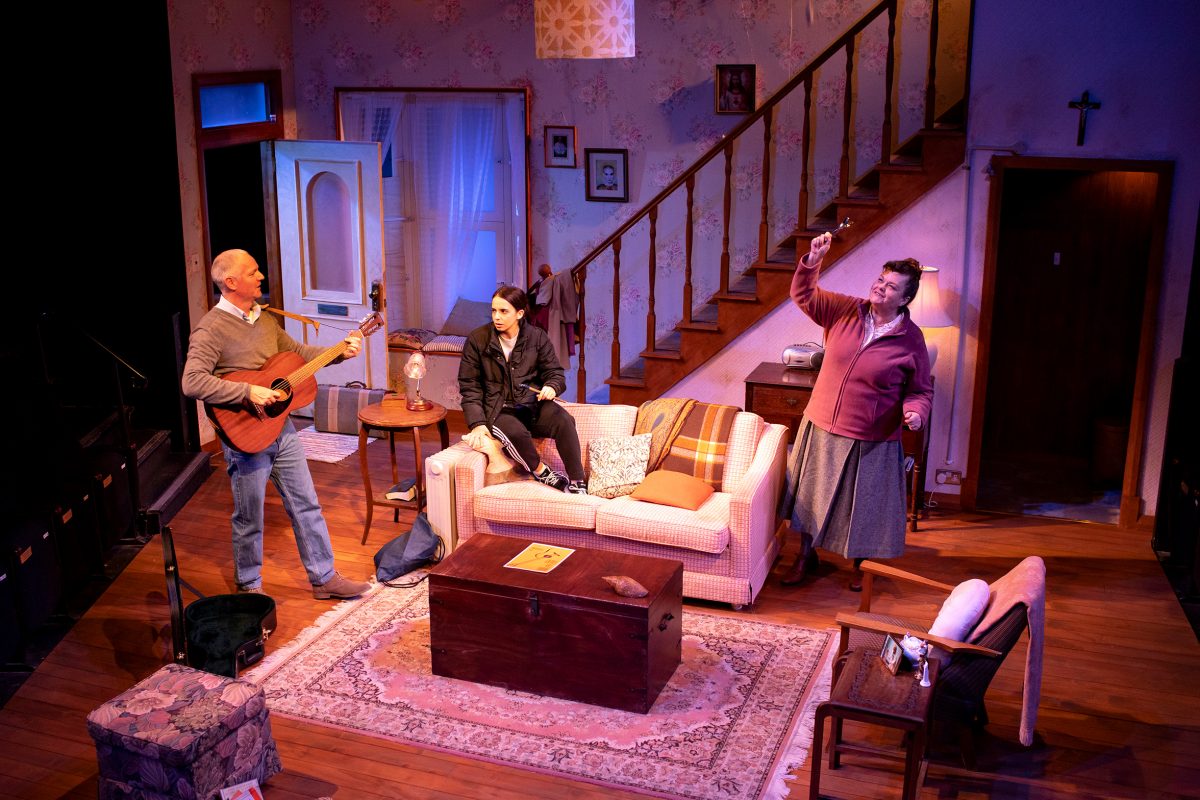 Three people are in a cosy living room set on stage. One man plays guitar, a woman sits on the sofa, and another woman stands nearby, raising her hand energetically. The room has warm lighting, stairs, and vintage décor.