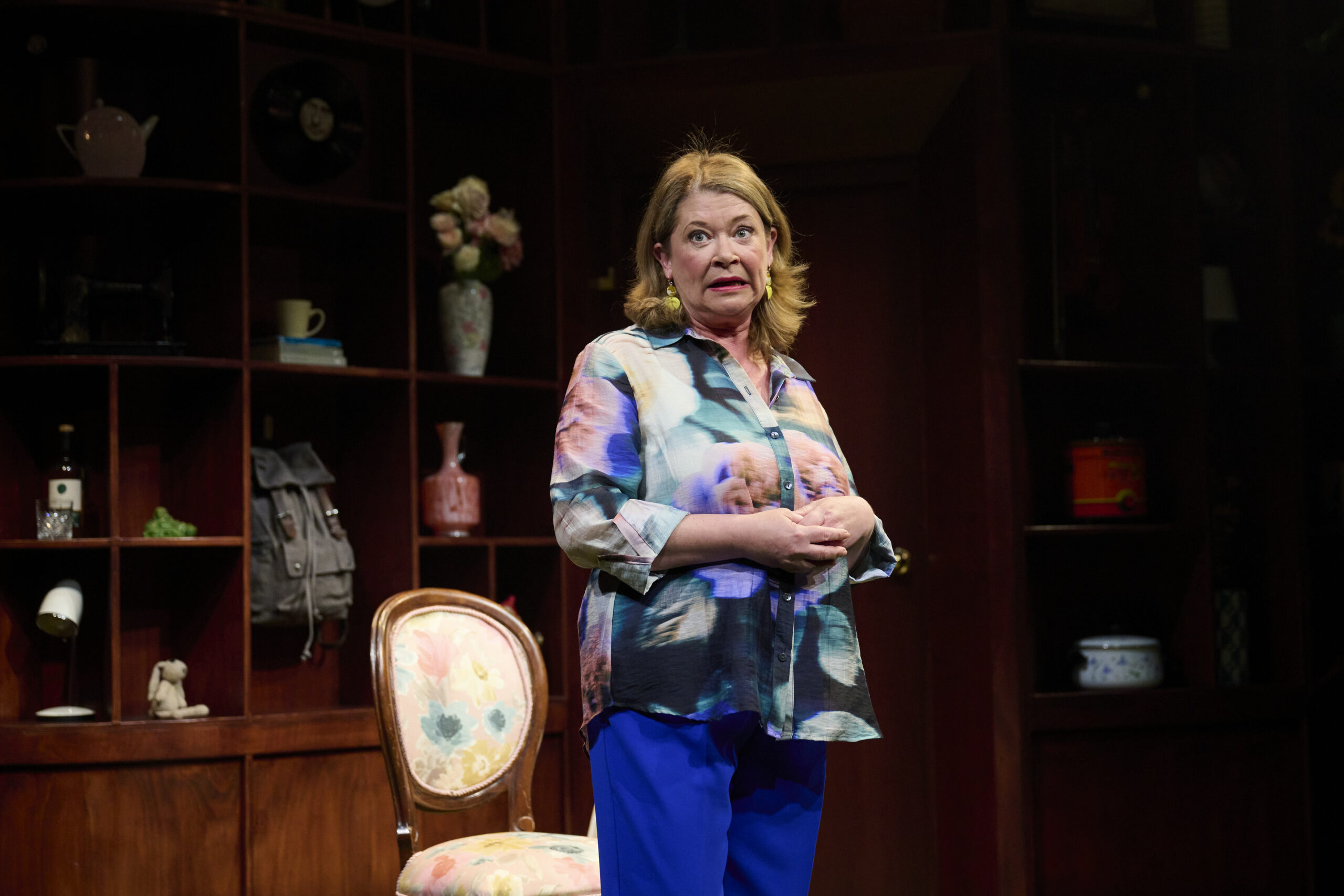 A woman stands on stage in front of wooden shelves filled with various objects. She wears a colourful blouse and blue trousers, with a patterned chair beside her, looking expressive as if speaking or performing.