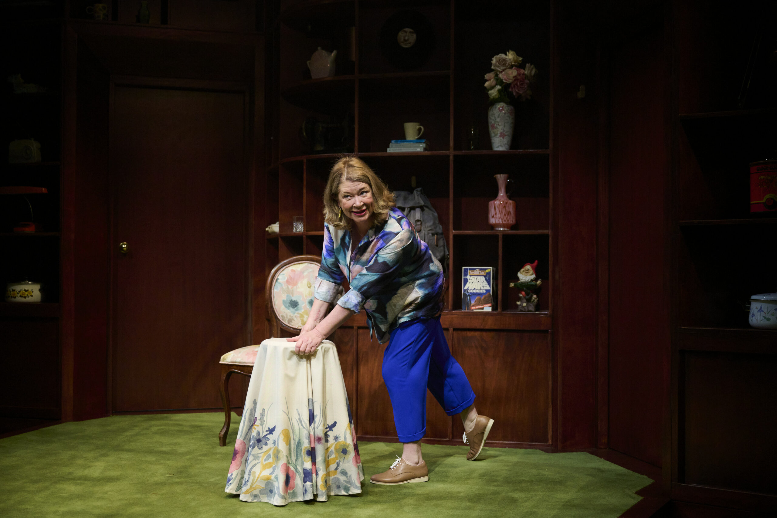 A woman with shoulder-length hair, wearing a blue blouse and trousers, leans forward with one hand on a chair covered by a floral cloth in a room with wooden shelves and decorative items.