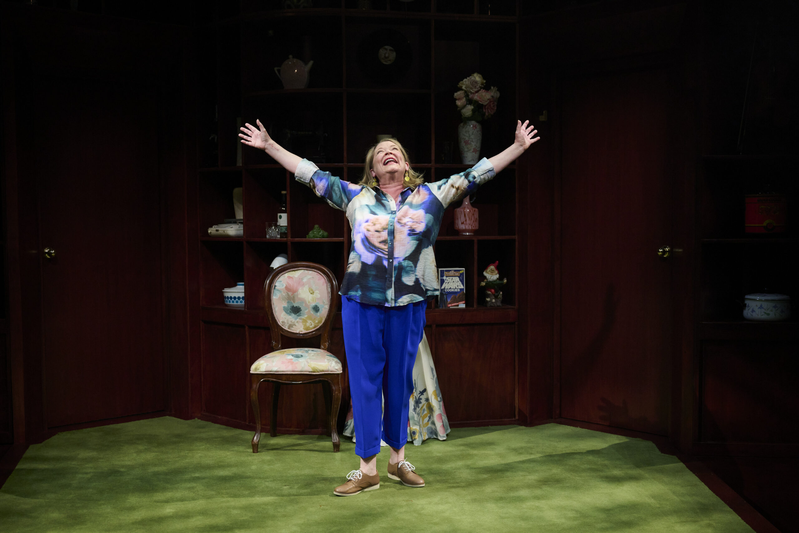 A woman stands on a green carpet with her arms raised and smiling, in front of wooden shelves filled with decorative items and a chair. She wears a floral shirt, blue trousers, and brown shoes.