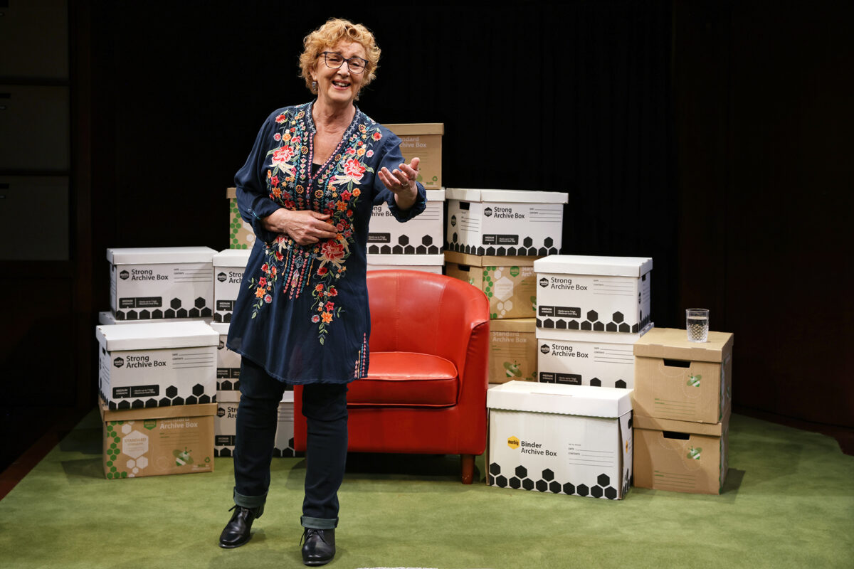 A woman with curly hair and glasses, wearing an embroidered dark dress, stands smiling and gesturing beside a red chair, surrounded by labelled archive boxes on a green carpet.
