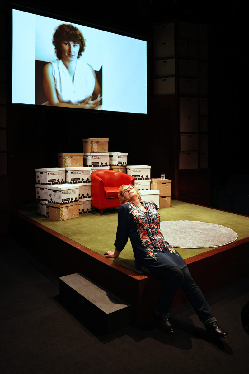 A person leans back dramatically at the edge of a stage, with an orange chair, stacks of document boxes, and a projected photo of a woman with curly hair and a white top in the background.