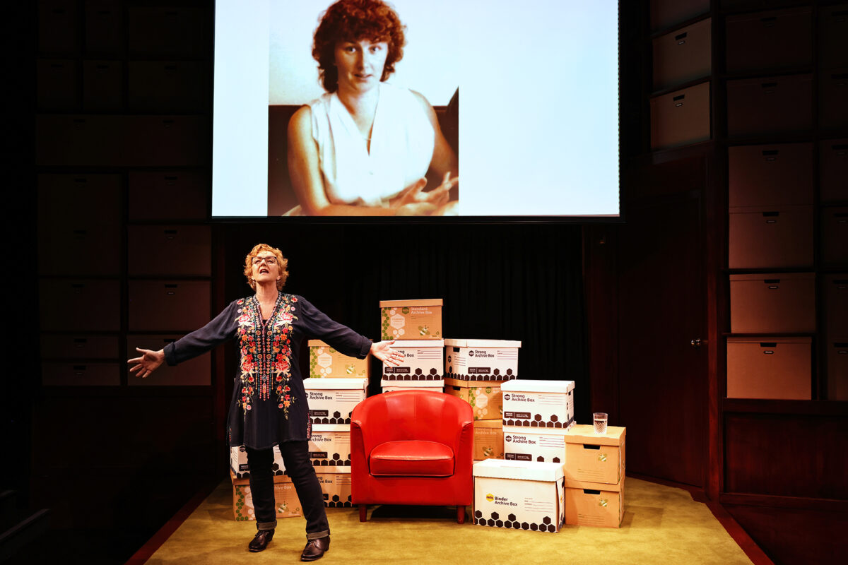 A woman stands on stage with arms outstretched in front of stacked filing boxes and a red chair, whilst a large projected photo of another woman appears behind her.