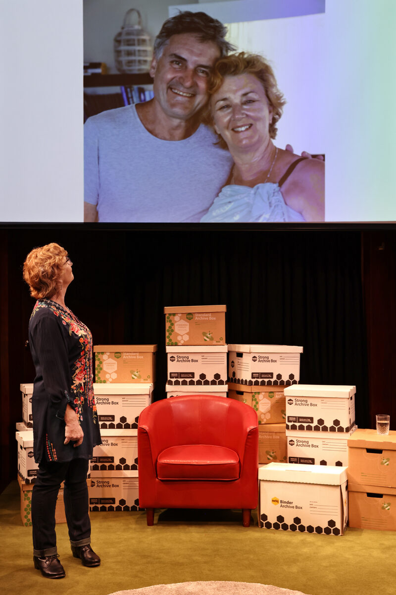 A woman stands on stage, looking up at a projected photo of a smiling couple. Behind her are stacked archive boxes, a red armchair, and a glass of water on a small table.